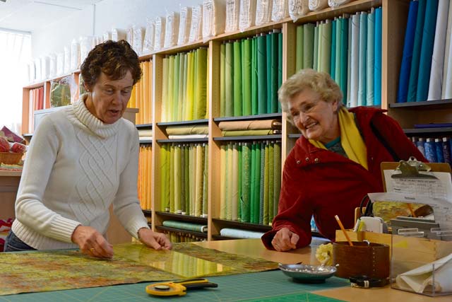 Sandy Vaughn, a volunteer at the new Kaiserslautern Arts and Cultural Center, measures a piece of fabric while talking with customer Ernie Gilgore.